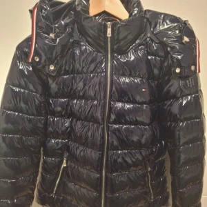 Mörkblå pufferjacka från Tommy Hilfiger - Säljer en snygg mörkblå pufferjacka från Tommy Hilfiger i mycket bra skick. Jackan har en glansig finish och dragkedja framtill. Perfekt för kyliga dagar med sin varma vaddering. Den har långa ärmar och en klassisk design med små detaljer i rött och vitt. Snygg glansig design✨