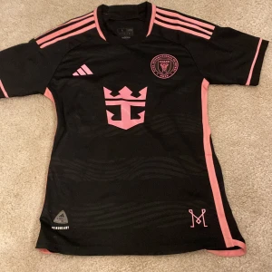 Svart och rosa fotbollströja - Säljer en snygg svart fotbollströja från Adidas med rosa detaljer och logga. Tröjan har korta ärmar och texten 'Freedom to Dream' på baksidan. Perfekt för fotbollsfans som vill sticka ut på planen! 🖤💖