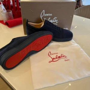 Marinblå sneakers från Christian Louboutin - Snygga marinblå sneakers från Christian Louboutin med den ikoniska röda sulan. Skorna har en stilren design med snörning och är tillverkade i mocka. Perfekta för att ge en touch av lyx till din outfit.