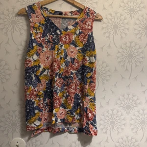 Blommig ärmlös topp - Säljer en färgglad ärmlös topp med blommigt mönster i rosa, blått och gult. Toppen har en v-ringad design och är perfekt för varma dagar.