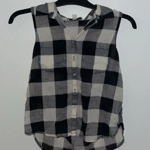 Svartvit rutig ärmlös blus - Snygg ärmlös blus i svartvitt rutmönster. Blusen har knappar framtill och en klassisk krage. Perfekt för en fin vardag .