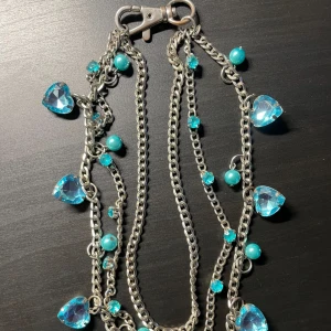 Silverfärgat halsband med blå detaljer - Charmigt silverfärgat halsband med blå hjärtformade stenar och pärlor. Kedjan är dekorerad med små blå kristaller som ger en glittrig effekt. Perfekt för att lägga till en färgklick till din outfit. 