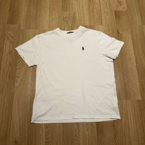 Vit t-shirt från Ralph Lauren - Säljer en klassisk vit t-shirt från Ralph Lauren med det ikoniska broderade logotypen på bröstet. T-shirten är kortärmad och tillverkad i mjuk bomull för en bekväm passform.