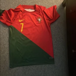 Portugal fotbollströja med nummer 7 - Säljer en Portugal fotbollströja från Nike i rött och grönt med gula detaljer. Tröjan har korta ärmar och nummer 7 på framsidan samt 'Ronaldo' på baksidan. 