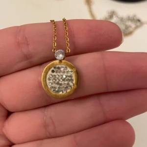 Guldhalsband med glittrig berlock - Elegant guldhalsband med en rund berlock fylld med glittrande stenar. Berlocken har en klar sten ovanför som ger extra bling. Perfekt för att lyfta vilken outfit som helst.