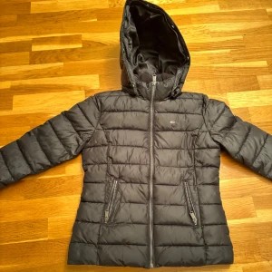 Svart dunjacka från Tommy Jeans - Säljer en svart dunjacka från Tommy Jeans med huva och dragkedja. Två praktiska fickor med dragkedja framtill.