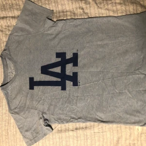 Grå t-shirt med LA-tryck - Säljer en grå t-shirt från Genuine Merchandise med stort LA-tryck i mörkblått på framsidan. Perfekt för sportiga tillfällen eller som en casual vardagströja. Tillverkad i Turkiet.