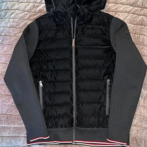 Unik Moncler cardigan - Säljer en snygg svart jacka/cardigan från Moncler med huva och dragkedja. Den har quiltade detaljer och ribbade muddar med röd och vit rand samt vaddering. Perfekt för kyligare dagar. Skriv för fler bilder!