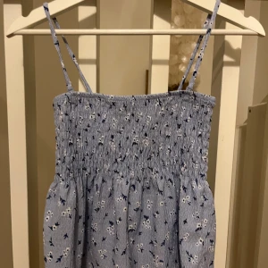 Blommig tröja från Gina Tricot - Söt blommig tröja från Gina Tricot i ljusblått med smala axelband. Perfekt för varma dagar med sitt luftiga och bekväma material. Den har ett elastiskt livstycke som ger en fin passform.