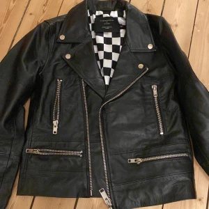 Svart skinnjacka med schackrutig insida - Snygg svart faux skinnjacka med schackrutig insida. Jackan har flera dragkedjor och knappar i silverfärg, vilket ger den en cool look. Perfekt för att ge din outfit en edgy touch. Jättebra skick!