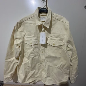 Light yellow Overshirt från A Day's March - Säljer en stilren light yellow overshirt från A Day's March. Skjortan är tillverkad i kraftig twill och har långa ärmar. Den har två bröstfickor med knappar och en klassisk krage. Perfekt för lager-på-lager-stil! Passar mig som är 183 cm 83 kg. 