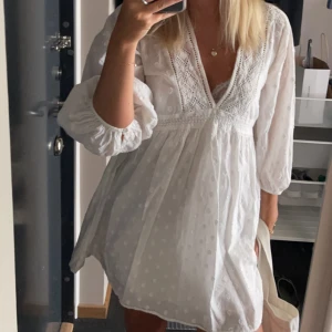 Vit klänning med spetsdetaljer - Säljer en söt vit klänning med spetsdetaljer och v-ringning. Klänningen har långa ärmar och ett prickigt mönster. Lite kort i modellen.
