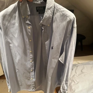 Blå skjorta från Polo Ralph Lauren - Snygg blå skjorta från Polo Ralph Lauren med klassisk krage och knappar framtill. Den har en broderad logga på bröstet och är långärmad. Perfekt för en stilren look.