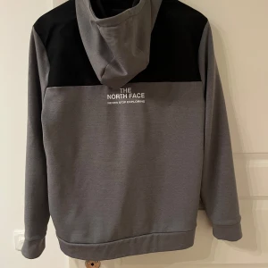 Grå och svart hoodie från The North Face - Säljer en snygg grå och svart hoodie från The North Face. Den har en dragkedja framtill och en stor logga på bröstet. Perfekt för kyliga dagar med sin bekväma passform och huva.