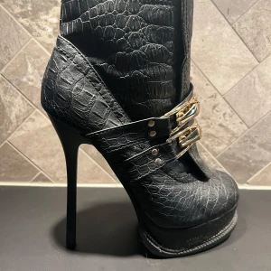 Svarta högklackade boots med krokodilmönster - Snygga svarta boots med hög klack och krokodilmönster. Skorna har dekorativa spännen i guld och en spetsig tå. Perfekta för att ge en edgy touch till din outfit. Dessa har varit en favorit hos mig, nu är det någon annans tur att ha kul i dessa! En omklackning behövs sen är det bara att ge sig ut på dansgolvet!