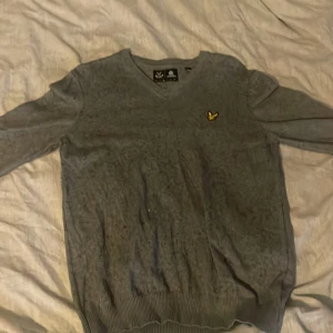 Grå tröja från Lyle & Scott - Säljer en stilren grå tröja från Lyle & Scott med deras ikoniska gula logga på bröstet. Tröjan har en v-ringad design och är perfekt för en avslappnad look. Passar bra till både jeans och chinos.