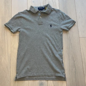 Grå pikétröja från Ralph Lauren - Säljer en stilren grå pikétröja från Ralph Lauren i slim fit. Tröjan har korta ärmar och en klassisk krage med två knappar. Den är dekorerad med den ikoniska broderade loggan på bröstet. Perfekt för en avslappnad men ändå snygg look som är perfekt till sommaren. hör av dig ifall du har några funderingar eller tankar👍