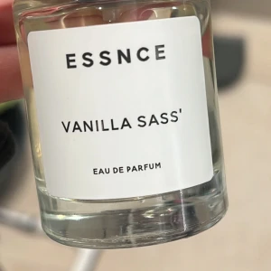 Essnce Vanilla Sass' Eau de Parfum - Säljer en elegant flaska med Essnce Vanilla Sass' Eau de Parfum. Doften har en söt och kryddig vaniljton som ger en självsäker och charmig känsla. Flaskan är genomskinlig med en stilren vit etikett.