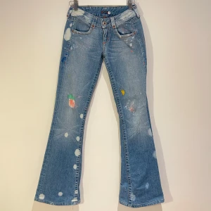 Blå bootcut jeans med färgstänk - Säljer ett par blå bootcut jeans med unika färgstänk i olika färger. Jeansen har en lågmidjad passform och klassiska femficksdetaljer. Perfekta för dig som vill ha en kreativ och avslappnad stil.  34,80cm