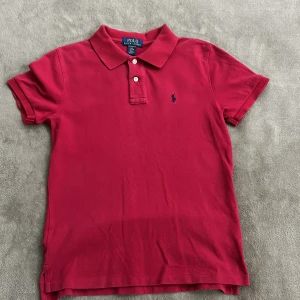 Röd pikétröja från Ralph Lauren - Säljer en klassisk röd pikétröja från Ralph Lauren med korta ärmar och knappar framtill. Tröjan har en broderad logotyp på bröstet och är perfekt för en stilren look. Passar bra till både jeans och chinos. 🌟