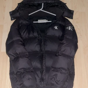 Svart pufferjacka från Calvin Klein - Snygg svart pufferjacka från Calvin Klein med dragkedja och logga på bröstet. Jackan har en hög krage och är perfekt för kyligare väder. Den är långärmad och har en stilren design.