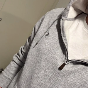 Grå hoodie från Ralph Lauren - Säljer en stilren grå hoodie från Ralph Lauren med dragkedja och broderad logga på bröstet. Tröjan har långa ärmar och en bekväm passform. Perfekt för en avslappnad look.