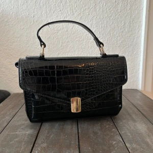 Svart krokodilpräglad handväska - Säljer en superfin svart handväska med krokodilpräglat mönster. Den har ett handtag och en justerbar axelrem för extra bekvämlighet. Väskan stängs med ett snyggt metallspänne och har gott om plats inuti för alla dina nödvändigheter. Perfekt för både vardag och fest!