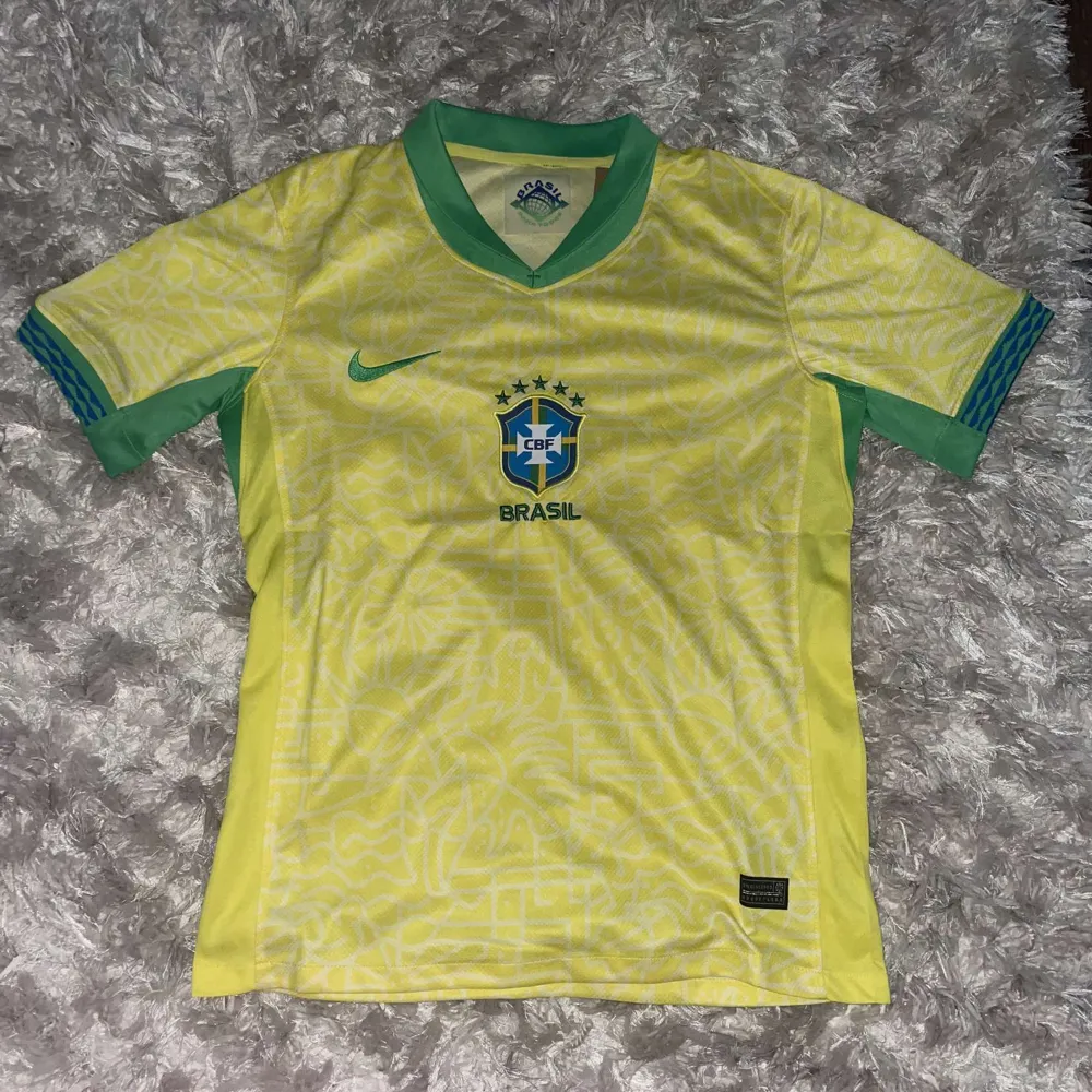 Säljer en gul Brasilien fotbollströja från Nike. Tröjan har korta ärmar och gröna detaljer vid kragen och ärmsluten. Den har Brasiliens emblem på bröstet och är i nyskick, fortfarande i förpackning.. T-shirts.