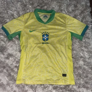 Gul Brasilien fotbollströja - Säljer en gul Brasilien fotbollströja från Nike. Tröjan har korta ärmar och gröna detaljer vid kragen och ärmsluten. Den har Brasiliens emblem på bröstet och är i nyskick, fortfarande i förpackning.