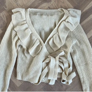 Beige stickad omlottkofta med volang - Säljer en superfin beige stickad omlottkofta med volangdetaljer. Den har en v-ringad design och långa ärmar, perfekt för höst och vår. Koftan är i ett mjukt och bekvämt material, perfekt att ha på sig när det är lite kyligare.