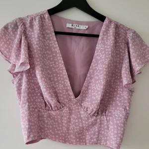 Ljuslila blommig topp från NA-KD - Säljer en superfin ljuslila topp från NA-KD med små vita blommor. Toppen har en djup v-ringning och korta ärmar med volangdetaljer. Resår i ryggen. Storlek: 36  Skickas inslagen i silkespapper i vattentätt paket.   Skicka pm för fler bilder. 