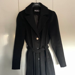 Svart kappa från Pieces - Säljer min fina svarta kappa från Pieces i mycket bra skick. Perfekt nu till höst och vinter, med knappar framtill för stängning. Passar både till vardags och festliga tillfällen!!