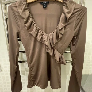Brun långärmad topp från Amisu - Säljer en snygg brun långärmad topp från Amisu i storlek S. Toppen har en v-ringad design med volangdetaljer längs halsringningen, vilket ger en elegant touch. Perfekt för både vardag och fest!