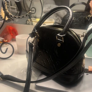 Svart handväska  - Säljer en elegant svart handväska, den är inte äkta men jättefin. Väskan har både handtag och en avtagbar axelrem, vilket gör den mångsidig och perfekt för både vardag och fest. Den är i mycket bra skick och har en dragkedja för säker förvaring.