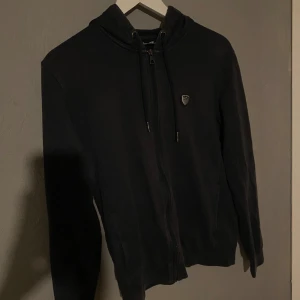 Svart kofta med dragkedja - Säljer en svart hoodie från EA Armani i bra skick. Den har en dragkedja framtill och en liten logga på bröstet. Perfekt för höst och vinter, och den är superbekväm att ha på sig. Passar både till vardags och chillkvällar. Hör av dig om du är intresserad! 🖤Tror storleken är S men passar Xs och M också. (PM OM DU VILL HA BÄTTRE BILDER)