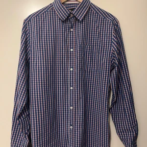 Rutig skjorta från Peak Performance  - Snygg rutig skjorta i blått och rött från Peak Performance. Perfekt för både vardag och fest! Skjortan har långa ärmar och knappar hela vägen ner. Den är i mycket bra skick och passar perfekt till höst och vår. Klassisk stil med en bröstficka för extra detalj.