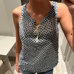 Mönstrad ärmlös topp från H&M - Säljer en superfin ärmlös topp med blått och vitt mönster från H&M . Toppen har en knytning framtill som ger en söt detalj. Perfekt för sommardagar eller en avslappnad vårlook. Materialet känns lätt och luftigt, vilket gör den bekväm att bära hela dagen. Jättebra skick, inga defekter!