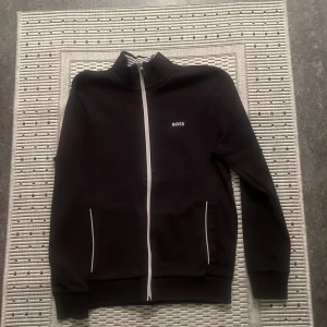 Svart tröja med dragkedja från BOSS - Säljer en stilren svart tröja från BOSS med vit dragkedja och broderad logga på bröstet. Tröjan har en hög krage med randigt mönster på insidan. Perfekt för både vardag och lite finare tillfällen. Passar bra till jeans eller chinos!