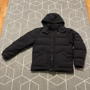 Svart dunjacka från Patagonia - Säljer en svart dunjacka från Patagonia i mycket bra skick. Jackan har en avtagbar huva och stängs med både dragkedja och knappar. Perfekt för kalla vinterdagar med sin pufferstil som håller dig varm. Storleken är herr M och den har en normal passform.