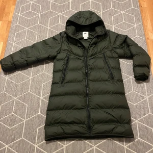 Svart dunjacka från Nike - Säljer en snygg och varm svart dunjacka som jag köpte från Nike för cirka 1 vecka sen. Perfekt för kalla vinterdagar med sin långa passform och huva. Jackan har en dragkedja framtill och är i mycket bra skick. Den är långärmad och har en stilren design som passar till det mesta. Perfekt för både vardag och utomhusaktiviteter!