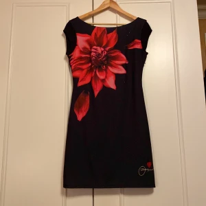Svart klänning från Desigual - Snygg svart klänning från Desigual med en stor röd blomma på framsidan. Den har en tight passform och är kortärmad, perfekt för festliga tillfällen. Klänningen är i mycket bra skick och har en unik design med broderad logga och hjärta.