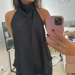 Svart ärmlös halterneck-topp - Snygg svart halterneck-topp i ett mjukt och flödande material. Perfekt för en utekväll eller fest. Den är ärmlös och har en elegant passform som ger en stilren look. Passar bra till både jeans och kjol. Köpt från bikbok! 