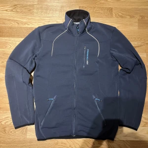 Mörkblå fleecejacka från Peak Performance - Säljer en mörkblå fleecejacka från Peak Performance i mycket bra skick. Jackan har en dragkedja framtill och två praktiska fickor med dragkedjor. Perfekt för kyliga höst- och vinterdagar. Materialet är 93% polyester och 7% elastan, vilket gör den både varm och stretchig. Passar perfekt för utomhusaktiviteter eller som ett extra lager under en skaljacka.