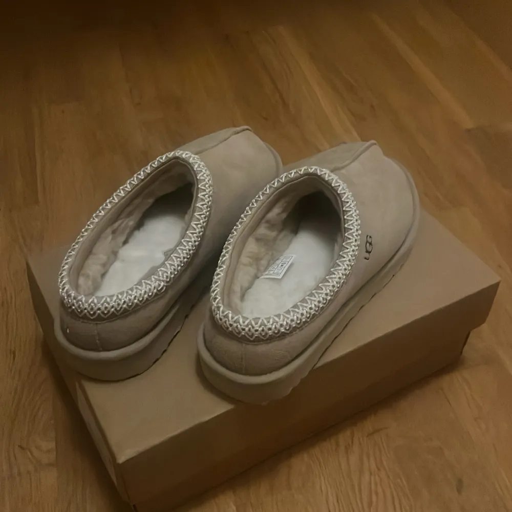 Mysiga beige tofflor från UGG i mycket bra skick. De är fodrade med mjuk päls och har en dekorativ kant runt öppningen. Perfekta för att hålla fötterna varma under kalla dagar. Säljes i originalkartong, aldrig använda.. Skor.