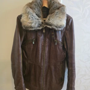 Brun skinnjacka med pälsdetaljer - Säljer en snygg brun skinnjacka med en fluffig pälsdetalj runt kragen. Jackan har en quiltad insida och dragkedja framtill. Perfekt för höst och vinter, håller dig varm och stilren. Passar både till vardags och festliga tillfällen.