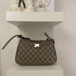 Mönstrad axelväska med dragkedja - Snygg axelväska i mycket bra skick med ett klassiskt mönster. Väskan har en praktisk dragkedja och justerbar axelrem. Perfekt för både vardag och fest!