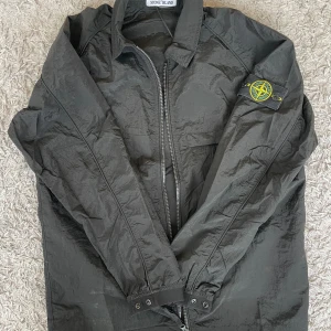 Grå jacka från Stone Island - Säljer en grå jacka från Stone Island i nytt skick. Den har en dragkedja framtill och en ikonisk logga på ärmen. Perfekt för höst och vår med sin stilrena design. Jackan är långärmad och gjord i ett lätt material som känns bekvämt att bära. Passar både till vardags och lite finare tillfällen.