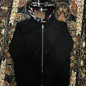 Burberry Hoodie - Säljer en helt ny och snygg svart hoodie från Burberry i storlek M. Den har en klassisk rutig insida i huvan och en dragkedja framtill. Perfekt för höst och vår, med en stilren look som funkar till det mesta. Huvan har dragsko för justerbar passform.