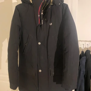 Svart parkajacka med huva - Säljer en stilren svart parkajacka från Fred Perry. Jackan har en praktisk huva och stängs med både dragkedja och knappar. Insidan är fodrad i beige för extra värme. Perfekt för kyliga höst- och vinterdagar. Passar både till vardags och lite finare tillfällen.
