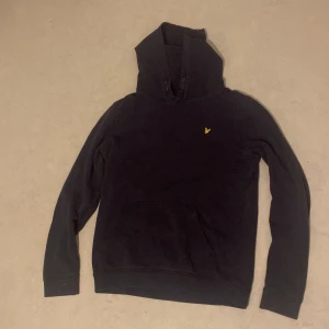 Mörk blå lyle & scott tröja - Säljer en mörklå lyle & scott hoodie med en liten gul logga på bröstet. Den har en klassisk känguruficka och är perfekt för både höst och vinter. Mysig och stilren, passar till det mesta. Hör av dig vid intresse!